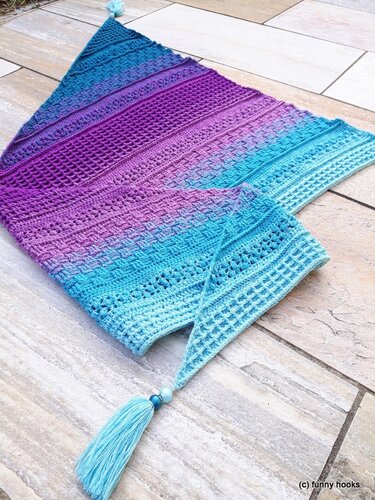 Crocheted triangle shawl laid flat showing aqua-to-purple ombre, textured and lacy stitch bands, and a beaded tassel.