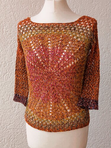 Gehäkeltes Damen‑Shirt in warmen Farben mit durchbrochenem Lochmuster, Boot‑Ausschnitt und Dreiviertelärmeln auf einer weißen Schneiderpuppe.