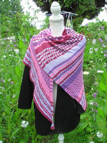 Gestricktes Dreieckstuch in Pink, Lila und Burgunder mit Streifen- und geometrischen Mustern auf einer Schaufensterpuppe vor Gartenpflanzen.