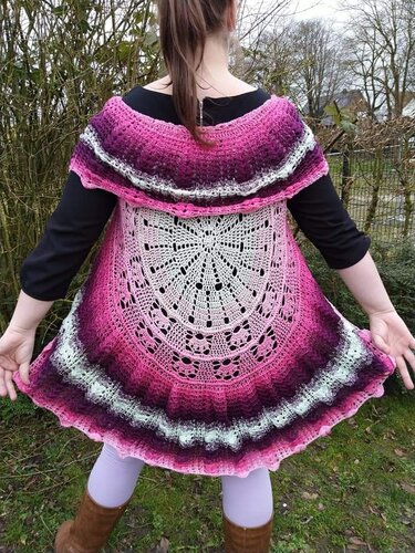 Person von hinten in einer pink-lila gehäkelten Weste mit rundem Mandala-Muster am Rücken und Rüschensaum.