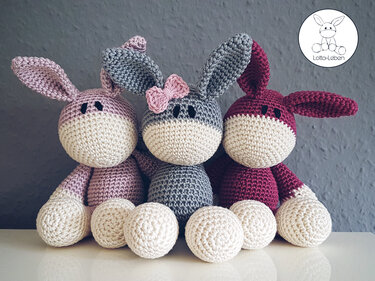 Three crocheted amigurumi bunnies in pink, grey, and burgundy with cream faces and round paws seated side by side; the grey bunny wears a small pink bow.
