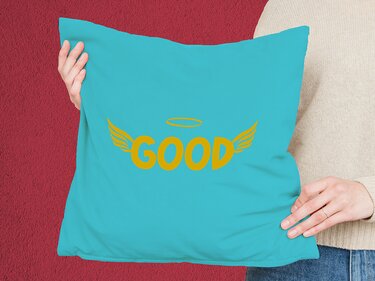 Türkisfarbenes Kissen mit gelbem Schriftzug „GOOD“, stilisierte Flügel und Heiligenschein, gehalten von Händen