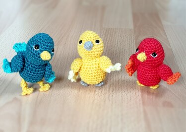 Drei gehäkelte Amigurumi-Vögel (blau, gelb, rot) auf Holzuntergrund