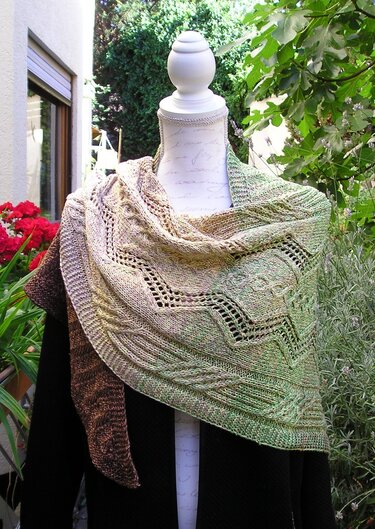 Dreieckiges, handgestricktes Tuch in Grün-, Beige- und Brauntönen mit Loch- und Zopfmuster auf einer Schneiderpuppe im Garten