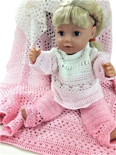 Babypuppe sitzt auf einer gehäkelten Decke und trägt ein weißes Oberteil und eine rosa Hose.