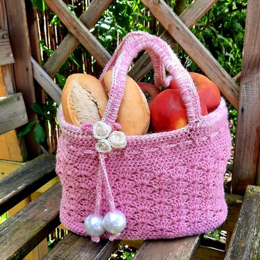 Rosa gehäkelte Tasche mit Henkeln, gefüllt mit Brötchen und Äpfeln, verziert mit Blüten und Perlen, auf einer Holzbank.
