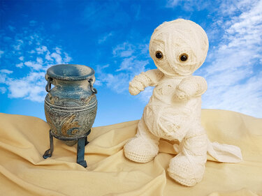 Beige gehäkelte Mumienpuppe mit dunklen Augen neben verzierter Vase vor blauem Himmel