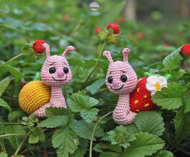 Zwei rosa gehäkelte Amigurumi-Schnecken mit gelbem und Erdbeerhaus zwischen Erdbeerpflanzen