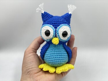 Hand-crocheted blue amigurumi owl with white eye rings, black round eyes, yellow beak and feet, held in a hand.