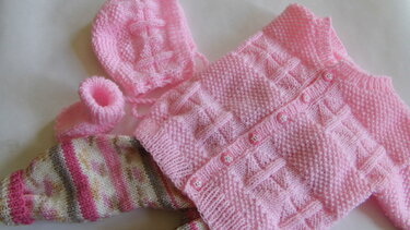 Gestricktes rosa Baby-Set mit Jacke mit Rautenmuster und sternförmigen Knöpfen, passender Mütze, kleinen Schühchen und gestreifter Hose.