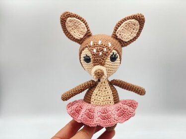 Crocheted amigurumi deer in brown and beige wearing a pink ruffled skirt, held by a hand.