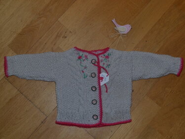 Hellgraue gestrickte Babyjacke mit rosa Zierkante und Blumenstickerei auf Holzboden