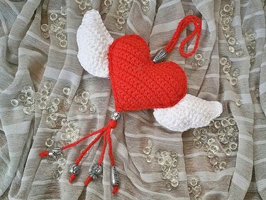 Gehäkeltes rotes Herz mit weißen Flügeln, roten Kordeln und silbernen Perlen auf hellem Tuch mit Glitzersteinen.