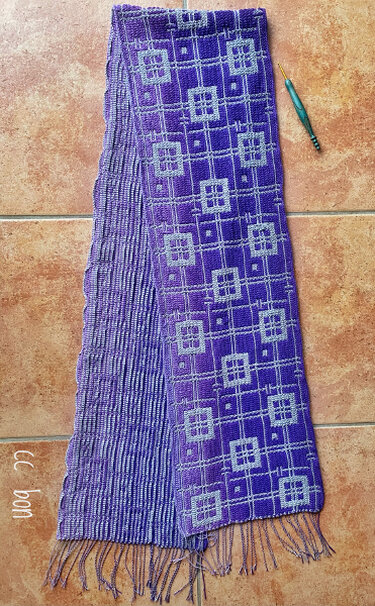 Long purple crocheted scarf with light square grid motifs, ribbed reverse and fringe; crochet hook beside it