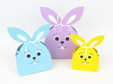 Drei bunte Hasen-Geschenkboxen aus Papier in Hellblau, Lila und Gelb mit weißen Schleifen