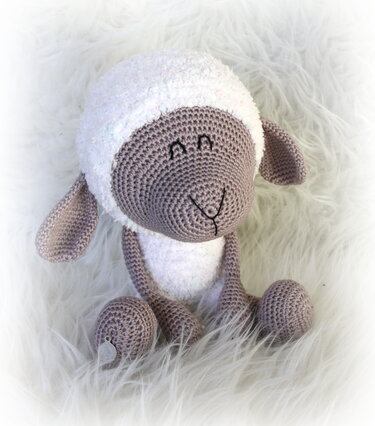 Gehäkeltes Amigurumi-Schaf mit weißem flauschigem Kopf und beige-braunem Gesicht, sitzend auf hellem Kunstfell