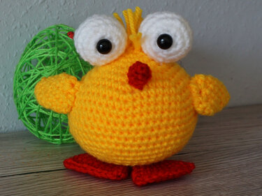 Round yellow crocheted chick amigurumi with large white eyes, red beak and red crocheted feet beside a green yarn egg