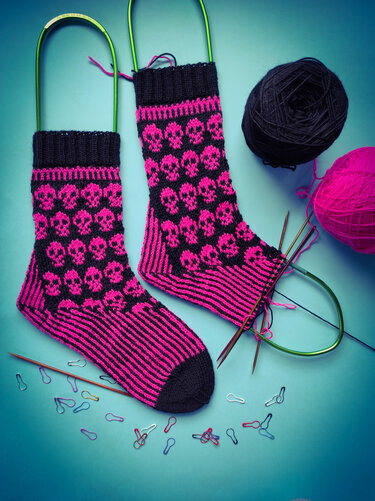 Unvollendete pink-schwarze Socken mit Totenkopfmuster auf Rundstricknadeln, daneben Garnknäuel und Maschenmarkierer
