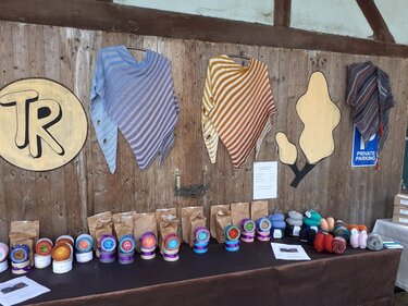 Drei gestreifte Dreieckstücher an einer Holzwand über einem Tisch mit Garnknäueln und Papiertüten.