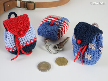 Close-up of three tiny crocheted backpack keychains with button flaps and drawstring cords beside keys and coins
