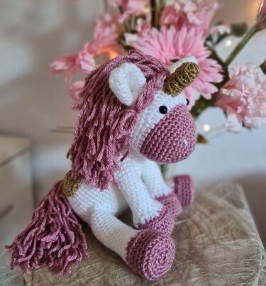 Gehäkeltes Einhorn weiß und rosa mit goldfarbenem Horn vor blumigem Hintergrund