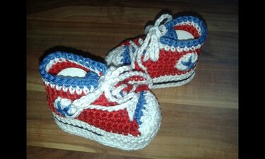 Nahaufnahme zweier gehäkelter Babyschuhe im Sneaker‑Stil mit Schnürbändern auf Holzuntergrund