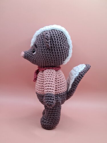 Side view of a crocheted amigurumi skunk wearing a pink sweater and red scarf on a pink background.