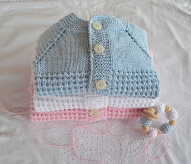 Stapel gestrickter Babyjacken in Pastell (Blau, Weiß, Rosa) mit sichtbaren Strukturmustern und Knopfleisten, dazu Beißring