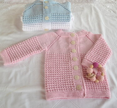 Rosa gestrickte Babyjacke mit Knöpfen und Beißring, gefaltete hellblaue Jacke im Hintergrund