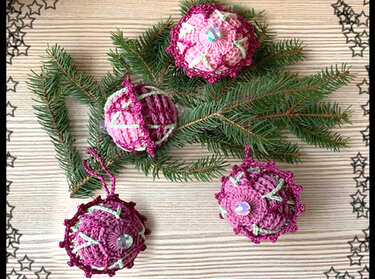 Vier gehäkelte Weihnachtskugeln in Rosa- und Burgundertönen mit dekorativen Mustern und Glitzersteinen auf Tannenzweigen