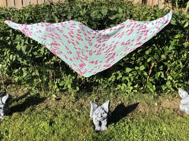 Gestricktes Dreieckstuch in Mint mit rosa Farbtupfen vor grüner Hecke, im Vordergrund kleine Gartenfiguren