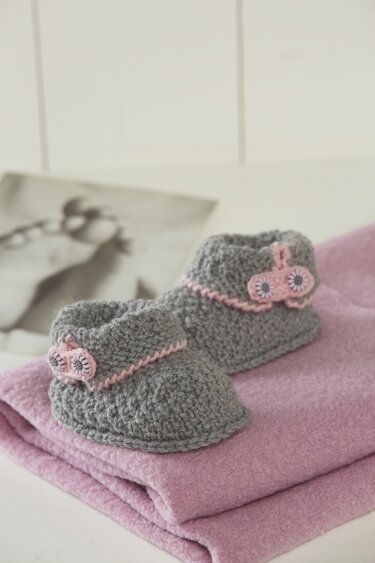 Grau gestrickte Babyschuhe mit rosa Kante und kleinen Blumen auf einer rosa Decke.