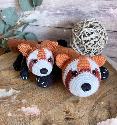 Zwei gehäkelte rote Panda-Schmusetücher mit weißen Gesichtern und schwarzen Pfoten auf Holztisch