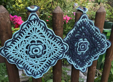 Zwei quadratische gehäkelte Topflappen in Hell- und Dunkelblau mit floralem Muster, an einem Holzzaun hängend