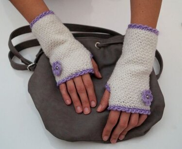Weiße gehäkelte fingerlose Handschuhe mit fliederfarbener Kante und kleiner Blüte auf grauer Tasche