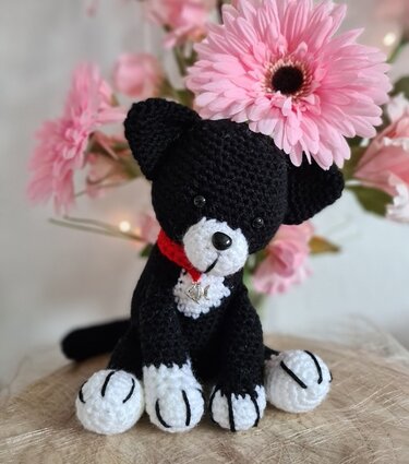 Schwarze gehäkelte Amigurumi-Katze mit weißen Pfoten und rotem Halsband vor rosa Blumen