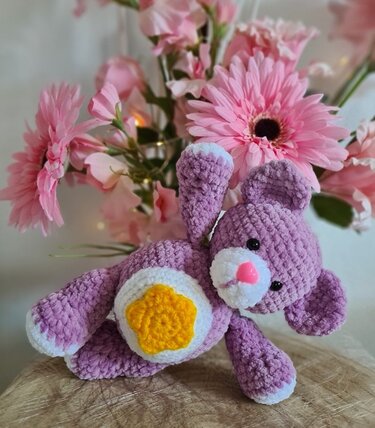 Gehäkelter lila Amigurumi-Teddy mit weißem Bauch und gelber Blüte vor rosa Blumen