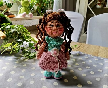 Gehäkelte Amigurumi-Puppe mit braunen Locken, mintgrünem Kragen und rosa Häkelrock mit Blüte