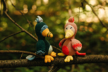 Zwei gehäkelte Amigurumi-Vögel in Grün und Rosa auf einem Ast vor unscharfem, grünem Hintergrund.