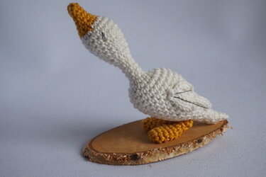 Gehäkelte weiße Gans mit orangefarbenem Schnabel und Füßen auf einer runden Holzscheibe