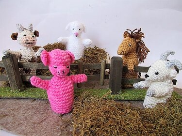Fünf gehäkelte Amigurumi-Fingerpuppen – rosa Schwein vorn, Kuh, Schaf, Pferd und Ziege vor einem Holzzaun