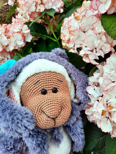 Nahaufnahme eines gehäkelten Amigurumi-Tieres mit flauschigem grauem Körper vor Hortensien.