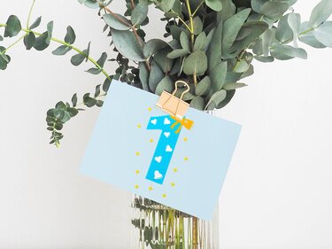 Hellblaue Karte mit großer Zahl 1, verziert mit weißen Herzen und gelben Sternen, an einer goldenen Klammer vor Eukalyptuszweigen