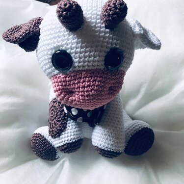 Gehäkelte Amigurumi-Kuh in Weiß und Braun mit rosa Schnauze und gepunktetem Halsband.