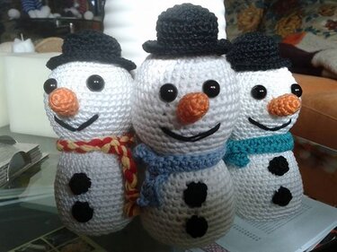 Nahaufnahme von drei weißen Amigurumi-Schneemännern mit schwarzen Hüten, Knopfaugen, orangefarbenen Nasen und bunten Schals.