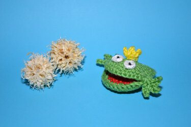 Kleiner gehäkelter Frosch mit rotem Maul und gelber Krone auf blauem Hintergrund neben zwei hellen Dekokugeln
