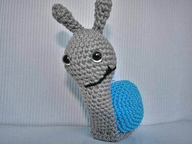 Gehäkelte graue Amigurumi‑Schnecke mit blauer Schale vor hellem Hintergrund
