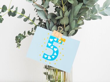 Karte mit blau dekorierter Zahl 5, Herz- und Sternmuster, befestigt mit goldener Binderklammer an einer Glasvase mit Eukalyptuszweigen