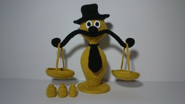 Gelbe und schwarze gehäkelte Amigurumi-Waage mit Zylinder, Schnurrbart, Krawatte, zwei Waagschalen und drei kleinen Gewichten.