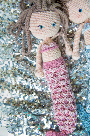 Gehäkelte Amigurumi-Meerjungfrau mit rosa Schwanzflosse, braunen Locken und blauen Augen auf glitzerndem Stoff.